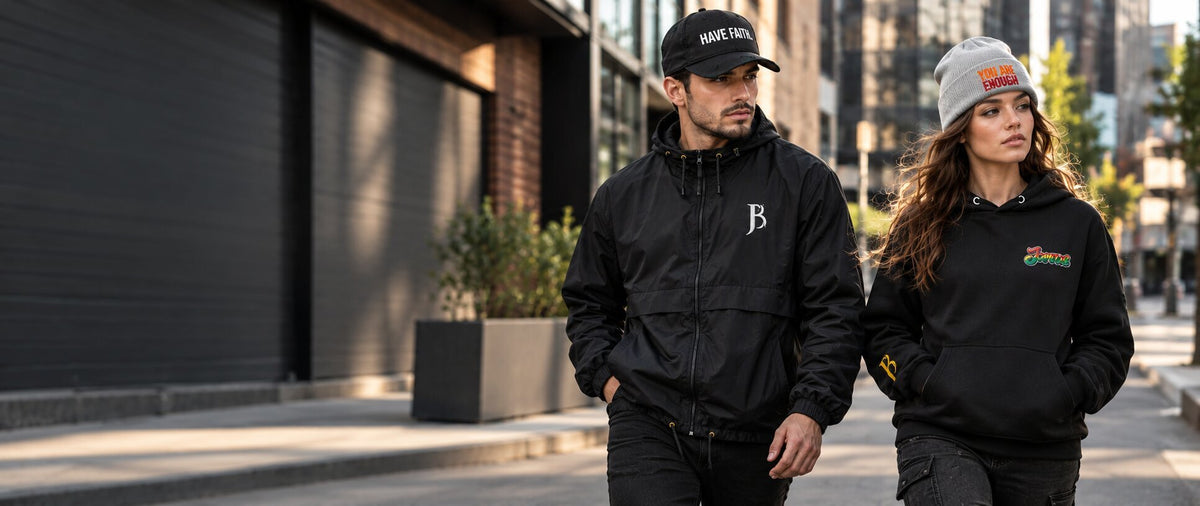 A man and a woman walking side-by-side on a city street. The man wears a black zip-up jacket with a white embroidered 'B' logo on the chest and a black cap.